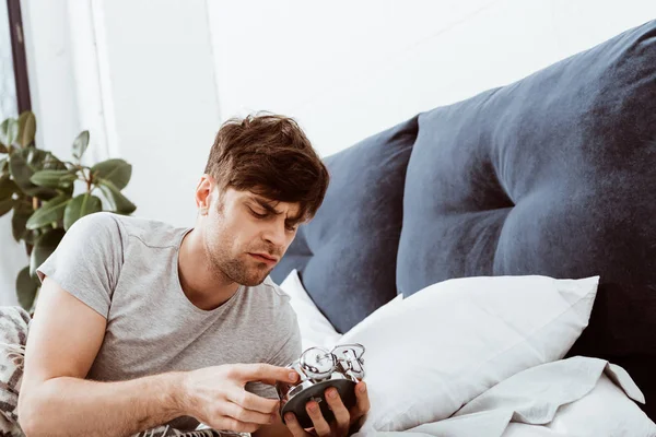 Irritated young man turning off alarm clock in bed at home — Stock Photo