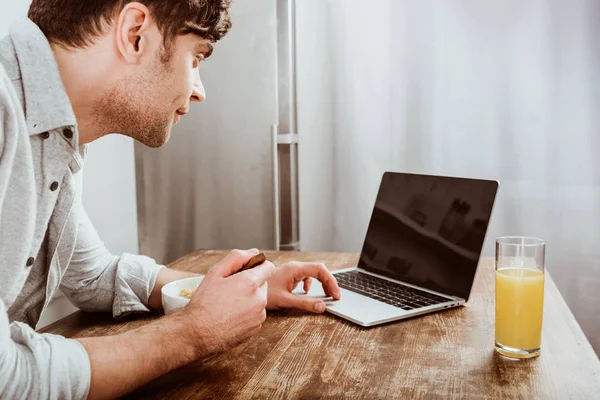 Vista lateral del freelancer masculino comiendo copos de maíz y trabajando en el ordenador portátil con pantalla en blanco en la mesa en la cocina — Stock Photo