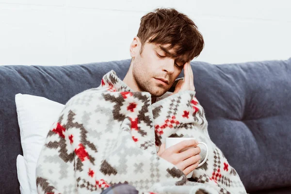 Sick man wrapped in blanket sitting with cup of tea in bed at home — Stock Photo