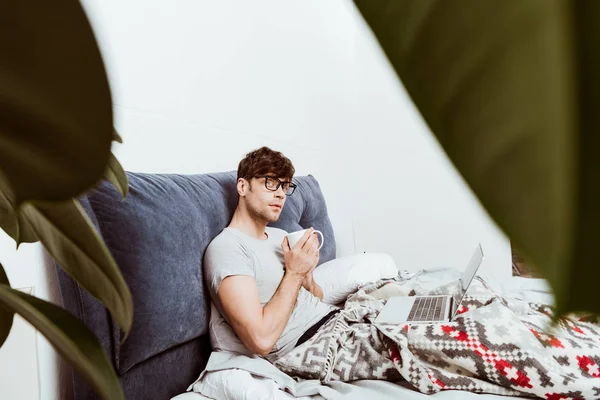 Foco seletivo de freelancer masculino pensativo com xícara de café sentado na cama com laptop em casa — Fotografia de Stock