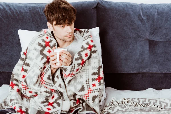 Vue grand angle de l'homme malade enveloppé dans la couverture assis avec une tasse de thé dans le lit à la maison — Photo de stock