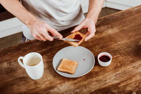 Visão parcial do homem espalhando torrada por geléia à mesa com xícara de café na cozinha em casa — Fotografia de Stock