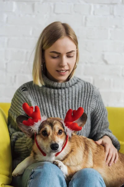 Glückliche junge Frau sitzt auf dem Sofa und trägt Corgi-Hund mit weihnachtlichen Hirschhörnern — Stockfoto