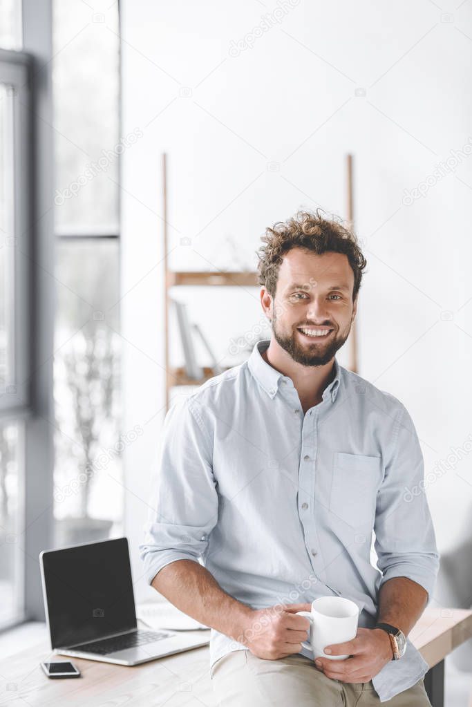 smiling businessman with cup of coffee sitting on table with laptop and smartphone in office