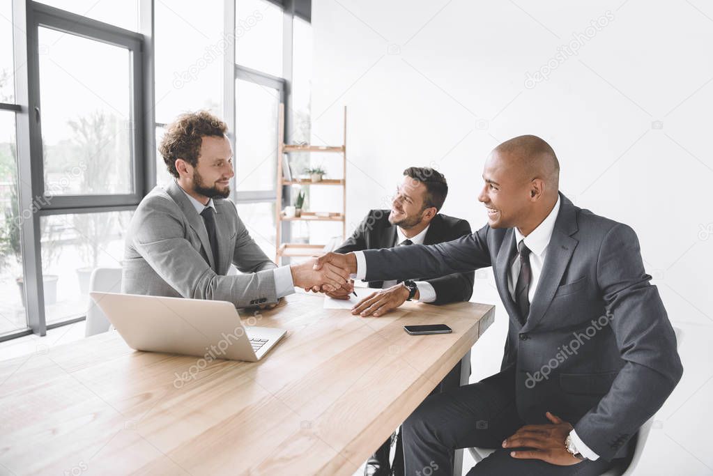 multiethnic smiling businessmen shaking hands at meeting at workplace with laptop