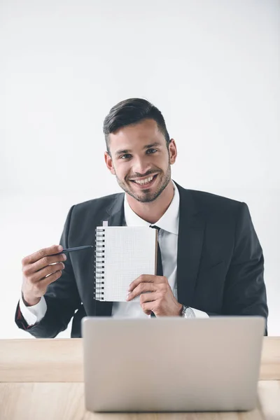 Enfoque selectivo del hombre de negocios sonriente apuntando a cuaderno en blanco en el lugar de trabajo aislado en blanco - foto de stock
