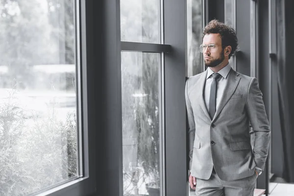 Young pensive businessman in suit looking out window in office — Stock Photo