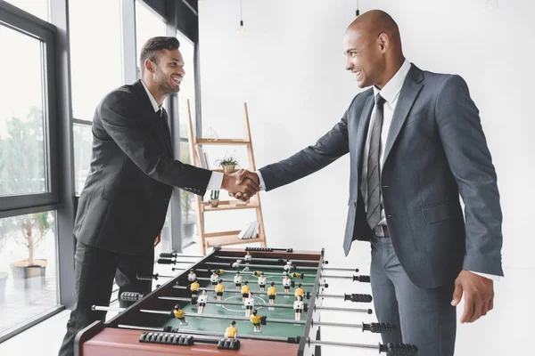 Vista lateral de homens de negócios sorridentes apertando as mãos depois de jogar futebol de mesa juntos — Fotografia de Stock