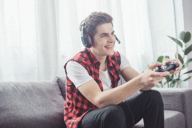 happy teenager with headset playing video game with joystick at home clipart