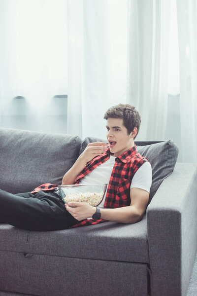 teen boy eating popcorn and watching tv on sofa at home