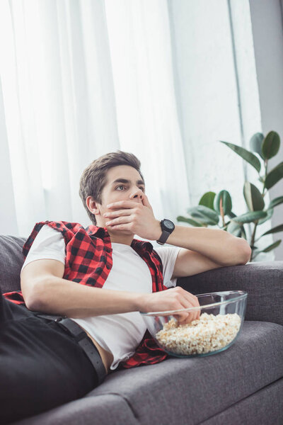 shocked teen boy with popcorn watching movie on sofa at home
