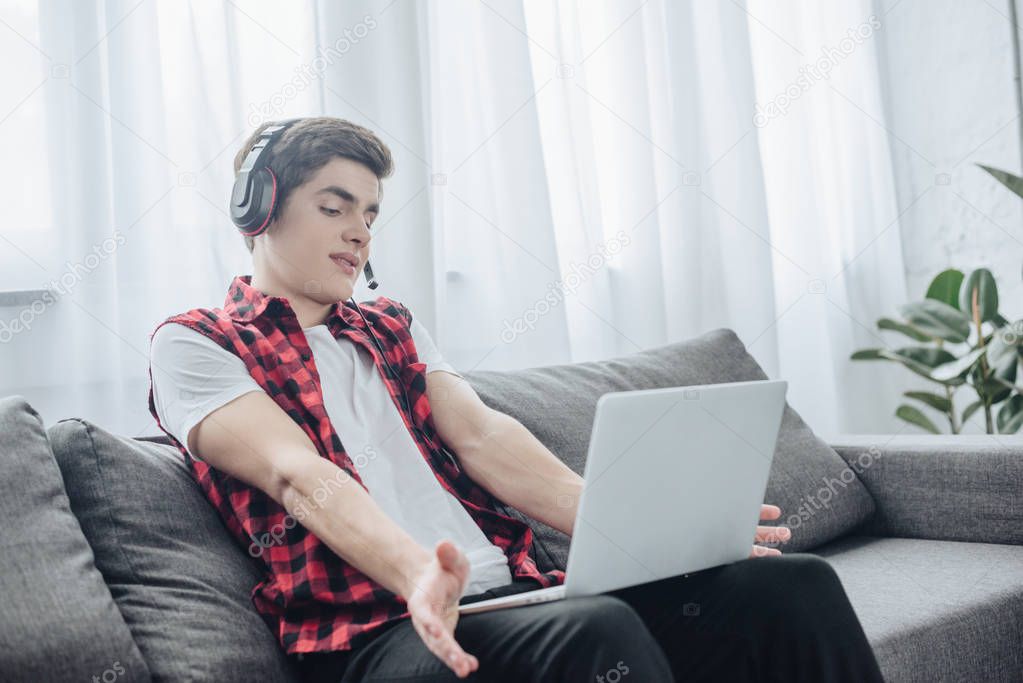 teen boy with headphones playing game on laptop while sitting on sofa 