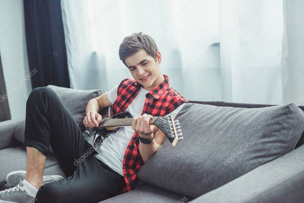 happy teenager playing electric guitar while sitting on sofa at home