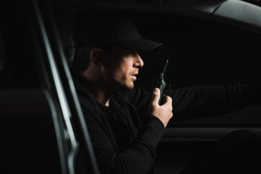 side view of male paparazzi in cap doing surveillance and using talkie walkie in his car clipart