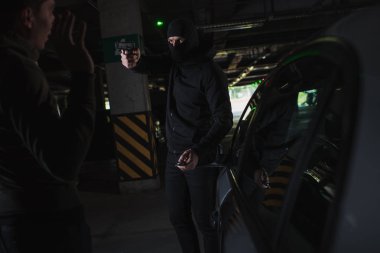 male mugger in balaclava aiming by gun on man standing near his car clipart