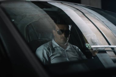 male undercover agent in sunglasses sitting in car with paper cup of coffee while doing surveillance  clipart
