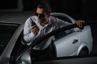 serious male undercover agent in sunglasses using talkie walkie near car  clipart