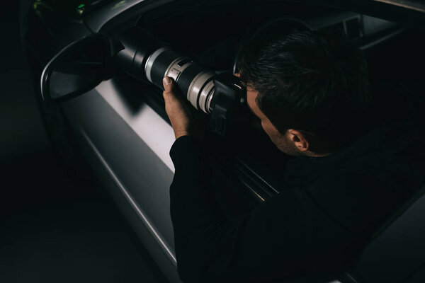 back view of undercover agent doing surveillance by camera with object glass from his car 