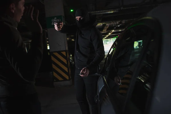 男子劫匪在巴拉克拉瓦瞄准枪对男子站在他的车附近 — 图库照片