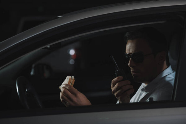 male undercover agent in sunglasses having lunch and using talkie walkie in car