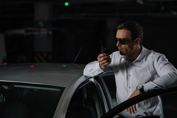 male undercover agent in sunglasses using talkie walkie near car 