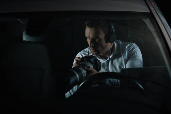 concentrated male private detective in headphones spying by camera with lens from car