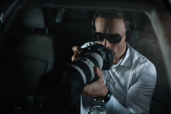 male private detective in headphones doing surveillance by camera with object glass from car