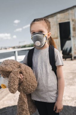 child in protective mask holding teddy bear on bridge, air pollution concept