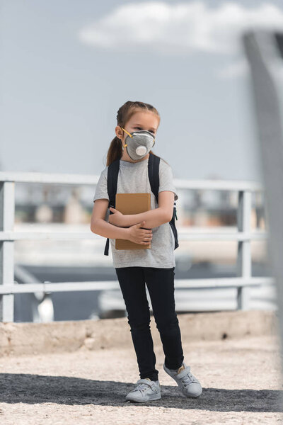 child in protective mask standing with bag and book on bridge, air pollution concept