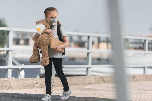 kid in protective mask walking with teddy bear and book on bridge, air pollution concept