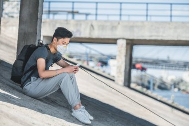 Köprünün altında oturan ve smartphone, hava kirliliği kavramı kullanarak koruyucu maske içinde Asya teen yan görünüm