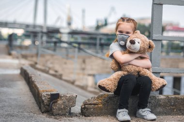 child in protective mask hugging teddy bear on street, air pollution concept clipart