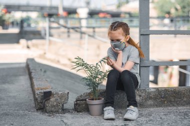 child in protective mask looking at green potted plant, air pollution concept clipart