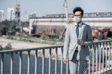 asian businessman in protective mask walking on bridge with coffee in paper cup, air pollution concept clipart