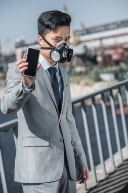 asian businessman in gas mask showing smartphone on bridge, air pollution concept clipart