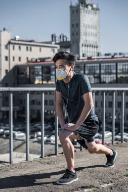 athletic asian teenager stretching in protective mask on bridge, air pollution concept clipart