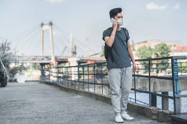 asian teen in protective mask talking by smartphone on bridge, air pollution concept