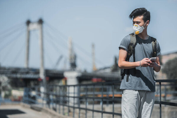 азиатский подросток в защитной маске держа смартфон и глядя в сторону, концепция загрязнения воздуха
