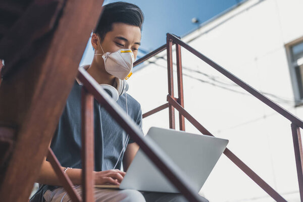 asian teen in protective mask using laptop on staircase, air pollution concept