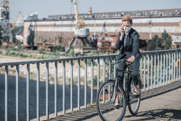 businesswoman in protective mask riding bicycle on bridge and talking by smartphone, air pollution concept