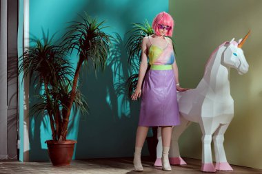attractive stylish girl in pink wig standing near unicorn and looking at camera clipart