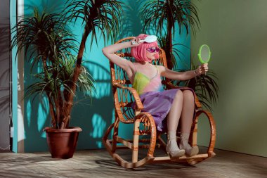 attractive girl in pink wig combing hair and looking at mirror while sitting in rocking chair clipart