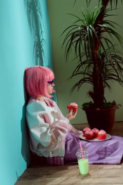 side view of stylish girl in pink wig eating cupcakes while sitting on floor clipart