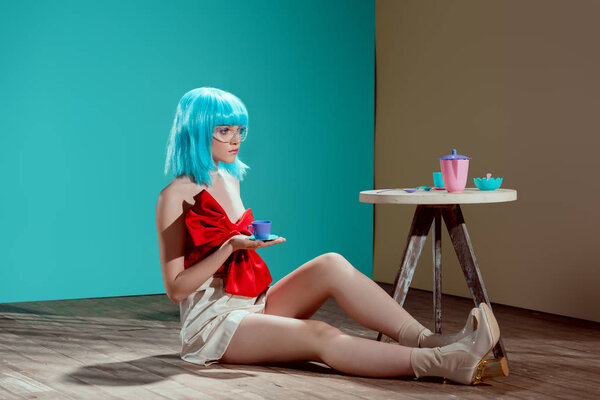 красивая модная девушка в синем парике, держа игрушечную чашку и отводя взгляд, сидя на полу
  