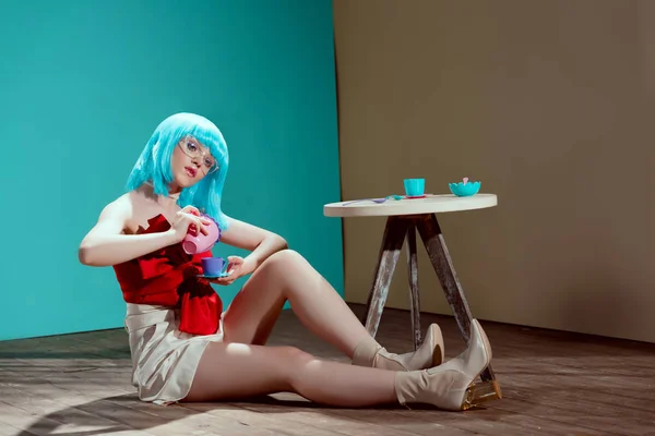 美丽的时尚女孩在蓝色假发举行玩具杯和茶壶 而坐在地板上 — 图库照片