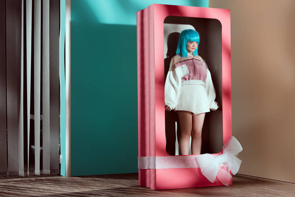 beautiful young woman in fashionable clothes and blue wig posing in decorative box with bow 