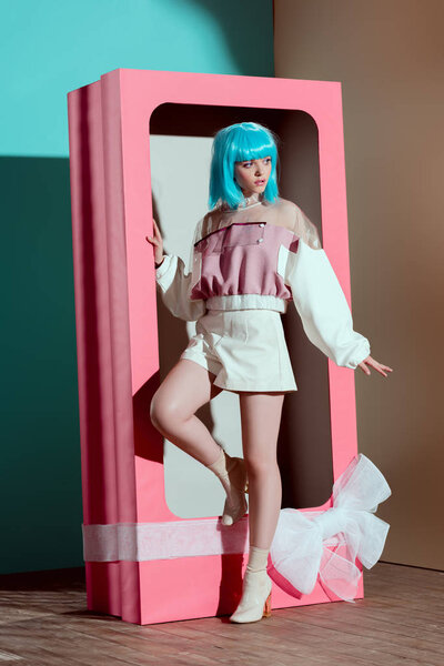 attractive fashionable girl in blue wig posing in decorative pink box with bow and looking away