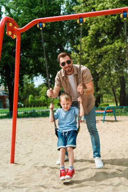 Baba ve oğul oyun park alanında, salıncakta eğleniyor ve kameraya bakıyor