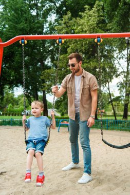 dad and son having fun on swing at playground in park clipart