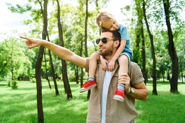 father pointing on something to son sitting on shoulders at park